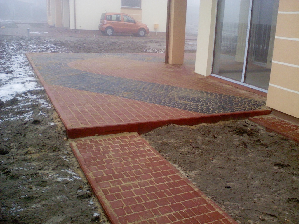 Ułożony taras z czerwonej kostki brukowej z czarnymi wstawkami, prowadzący do budynku z dużym oknem tarasowym, widoczna ścieżka z kostki i błotniste podłoże, w tle zaparkowany samochód w pochmurny...